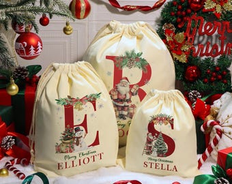 Puede incluir: Tres bolsas de regalo navideñas de color crema con letras rojas e ilustraciones festivas. Las bolsas presentan las letras "E", "R" y "S", con los nombres "Elliott" y "Stella" impresos debajo. Decoraciones navideñas rodean las bolsas.