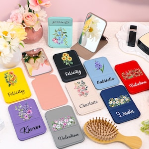Könnte beinhalten: Eine Sammlung personalisierter Telefonständer in verschiedenen Farben, jeweils mit floralen Designs und Namen verziert. Die Ständer zeigen Sonnenblumen, Lavendel und andere Blumen. Zu den Farben gehören Gelb, Schwarz, Blau, Rot und Rosa. Eine Holzbürste ist ebenfalls vorhanden.