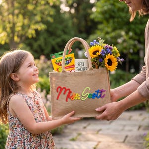 May include: A tan burlap tote bag featuring the name Miss Scott in colorful, textured script lettering. The bag contains a box of Crayola crayons, a white mug with the text BEST TEACHER, and a bouquet of sunflowers and blue wildflowers.