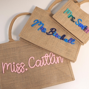 May include: Three rectangular jute tote bags with rounded handles. Each bag features personalized text in raised, cursive yarn lettering. The bags display the names "Mrs. Smith" in teal and blue, "Mrs. Birchall" in blue and purple, and "miss. Caitlin" in light pink.