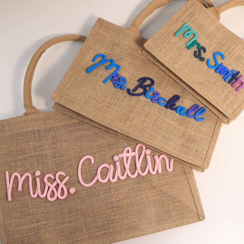 May include: Three rectangular jute tote bags with rounded handles. Each bag features personalized text in raised, cursive yarn lettering. The bags display the names "Mrs. Smith" in teal and blue, "Mrs. Birchall" in blue and purple, and "miss. Caitlin" in light pink.