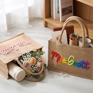 May include: Two rectangular jute tote bags with sturdy handles. One bag features the pink embroidered text 'Miss Caitlin' and contains a bouquet of roses and a white tumbler. The other bag features the multicolored embroidered text 'Mrs Scott'.