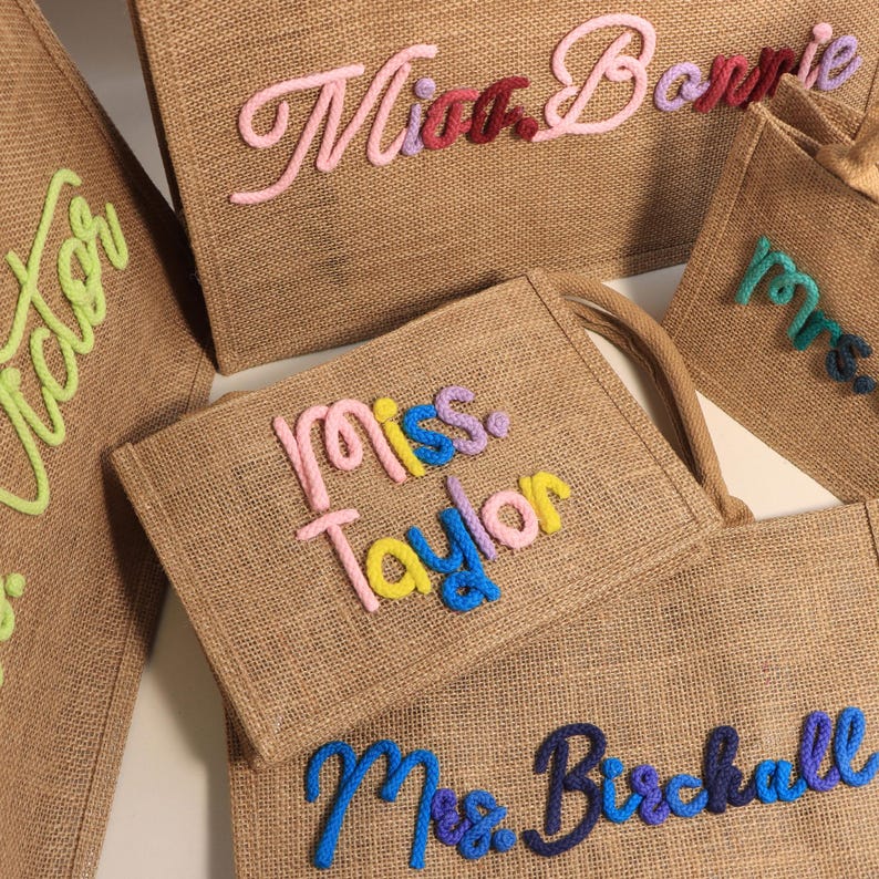 May include: A collection of tan jute tote bags featuring personalized names and titles in raised, colorful yarn lettering. The bags display various names including Miss. Bonnie, miss. Taylor, and Mrs. Birchall, with text in pink, yellow, blue, and green.