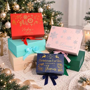 Op de afbeelding: Een collectie rechthoekige geschenkdozen met lintsluiting in diverse kleuren zoals rood, lichtblauw, beige, roze, donkergroen en marineblauw, elk gepersonaliseerd met namen en feestelijke afbeeldingen zoals sneeuwvlokken en ornamenten.