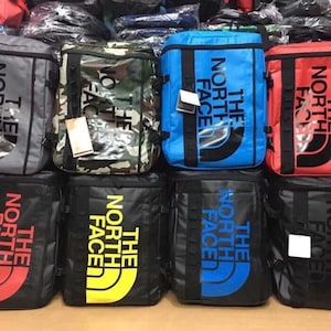 May include: Eight different The North Face brand backpacks in various colours, including black, grey, blue, red, and camouflage. The backpacks have the brand name and logo printed on the front.