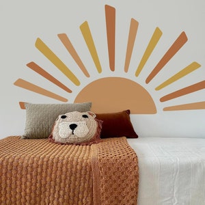 May include: A white bed with a brown crocheted blanket and two pillows. One pillow is a crocheted lion head and the other is a brown pillow. The wall behind the bed has a large painted sun with orange, yellow, and brown rays.
