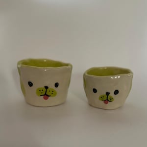 Puede incluir: Dos pequeñas macetas de cerámica con forma de cara de perro. Las macetas son verdes y blancas con ojos negros y una lengua rosa.