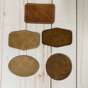 May include: Five leather patches in different shapes and sizes. The patches are brown and have a stitched edge. The patches are on a white wooden background.