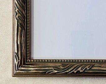 May include: Close-up of a decorative picture frame with a detailed, ornate design. The frame is a warm, antique gold color with carved leaf and scroll details. The frame surrounds a white, blank space, ready for a photo or artwork.