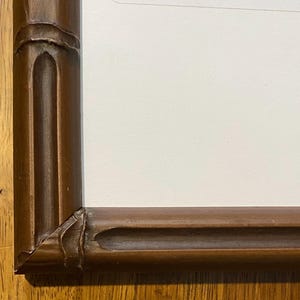 May include: A close-up of a brown wooden picture frame with a bamboo-like design. The frame has a rectangular shape with rounded corners and a light-colored interior. The frame is sitting on a wooden surface.
