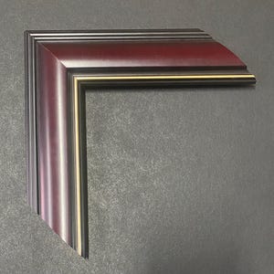 May include: A corner section of a picture frame with a rich burgundy finish. The frame has a layered design with a thin gold accent line. The wood grain is visible, adding texture. The frame is set against a dark gray background.