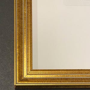 May include: Close-up of a gold-colored picture frame with a textured surface. The frame has a layered design and a bright, reflective finish. The frame surrounds a white surface.
