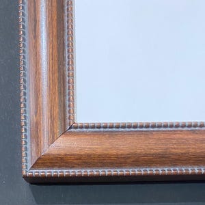 May include: A rectangular wooden picture frame with a warm brown finish. The frame has a detailed, textured design along the edges and corners, and a white interior. The frame is designed to hold a photograph or artwork.