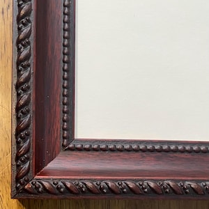 May include: Close-up of a dark red picture frame with a decorative rope-like border. The frame has a beveled edge and a cream-colored matting. The corner of the frame is visible, showing the joinery.