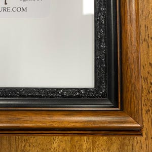 May include: Close-up of a framed artwork. The frame is made of dark brown wood with a black inner border. The frame has a decorative, ornate design. The artwork inside is white.