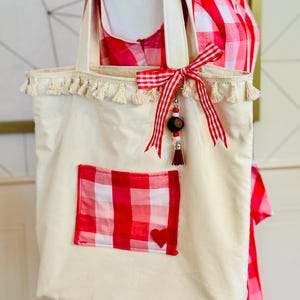 May include: Cream-colored tote bag with a red and white gingham pocket and a small red heart. The bag has cream-colored handles, a tassel trim, and a red and white ribbon bow with a decorative charm. The bag is made of a sturdy fabric.