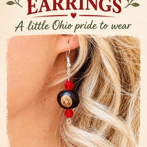 May include: Handmade Buckeye earrings featuring a dark brown, round nut with a light tan center, accented by two faceted red beads on a silver-toned metal hook. The text reads HANDMADE BUCKEYE EARRINGS A little Ohio pride to wear.