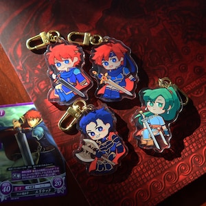 Puede incluir: Cinco llaveros acrílicos con personajes de dibujos animados con espadas y hachas. Los personajes tienen pelo rojo, azul y verde, y visten trajes azules y rojos. Cada llavero tiene un cierre dorado. Una tarjeta con texto japonés está en el fondo.