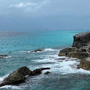 May include: A rocky coastline with turquoise water crashing against the shore. The water is clear and blue, and the sky is cloudy.