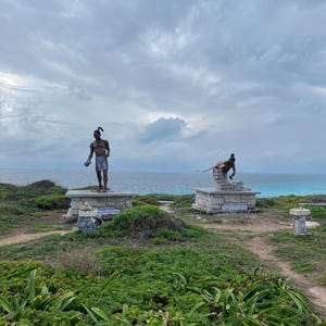 May include: Two statues of people in traditional clothing stand on stone platforms overlooking a turquoise ocean. The sky is cloudy and the beach is sandy.