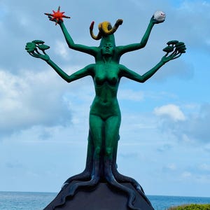 May include: A large, green statue of a woman with multiple arms, holding various objects. The statue has a tree-like base and is set against a backdrop of the ocean and sky. The statue's arms hold a star, a snake, a sphere, and two small trees.