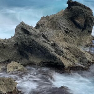 May include: A rocky cliff face with waves crashing against it. The water is a light blue and the rocks are a dark gray. The waves are blurred due to the long exposure.