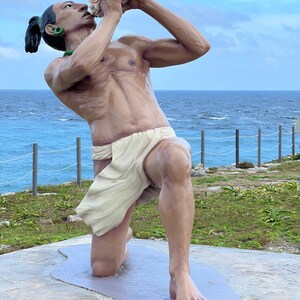 May include: A statue of a man kneeling and blowing a conch shell. The statue is made of brown and white materials and is set against a backdrop of blue water and a cloudy sky.