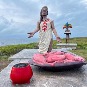 May include: Outdoor sculpture featuring a large statue of a woman in a white dress with red accents, arms outstretched. A smaller statue of a man with a colorful headdress stands nearby. A red bowl and a large, pink, sculpted form are in the foreground.