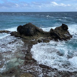 May include: A coastal scene featuring a rocky island in the ocean. The water is a deep blue-green, with white waves crashing against the rocks. The sky is overcast with a mix of blue and gray clouds.