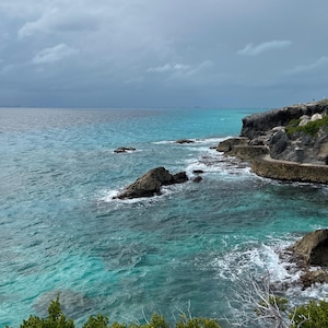 May include: A rocky coastline with turquoise water crashing against the shore. The sky is overcast with gray clouds.