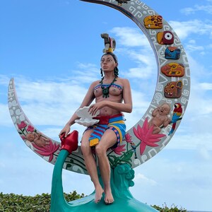 May include: A colorful sculpture of a woman sitting on a crescent moon. The woman is wearing a headdress and a skirt with intricate designs. The moon is decorated with flowers, birds, and other symbols. The sculpture is set against a blue sky with white clouds.