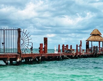 May include: A wooden pier extends over turquoise water, leading to a thatched roof gazebo. A black metal gate with decorative accents is at the beginning of the pier.