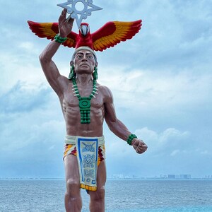 May include: A statue of a man wearing a feathered headdress and a green beaded necklace, holding a white star-shaped object above his head. The statue is standing on a grassy area with a body of water in the background.