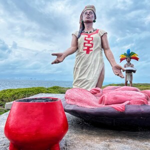 May include: A sculpture of a woman in a white dress with red accents, standing on a platform with a large, red bowl. The woman is holding out her hand, and there is a smaller sculpture of a person in a colorful headdress holding a plate in front of her.