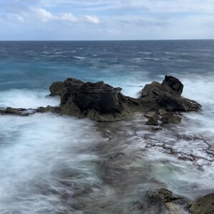 May include: A rocky outcropping juts out into the ocean, with waves crashing against the rocks and creating a misty, ethereal effect. The water is a deep blue, and the sky is a light blue with wispy clouds.