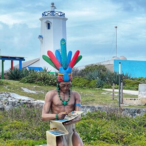May include: A statue of a person wearing a colorful feathered headdress and holding a book. The statue is kneeling and is in front of a white lighthouse.