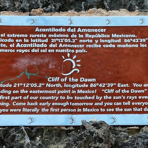 May include: A brown sign with white text reads "Acantilado del Amanecer" and "Cliff of the Dawn". The sign explains that this is the easternmost point in Mexico and that it receives the first rays of the sun each morning. The sign is attached to a rocky surface.