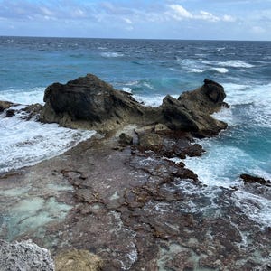 May include: A coastal scene featuring a rocky shoreline with dark, weathered rocks and a turquoise ocean. Waves crash against the rocks, creating white foam. The sky is overcast with a mix of blue and gray.