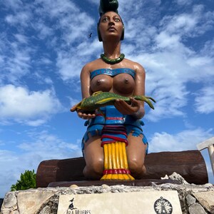 May include: A statue of Ixchel, the Mayan goddess, is depicted kneeling and holding a fish. The statue has a green headdress, a blue and red outfit, and a green necklace. The statue is on a stone base with text in Spanish and the words "Isla Mujeres".