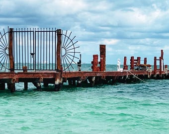 May include: A wooden pier with a black metal gate at the entrance extends over turquoise water. A thatched roof gazebo is at the end of the pier.