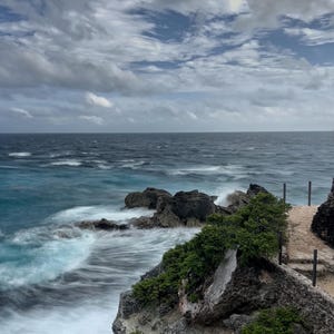 May include: A rocky cliff overlooking a blue ocean with white waves crashing against the shore. The sky is cloudy with a mix of white and gray clouds.