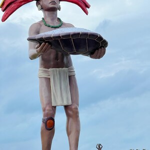 May include: A large statue of a person wearing a colorful headdress and holding a large turtle shell filled with white sand. The statue is standing on a concrete platform with a smaller statue of a person in the background.