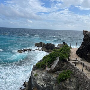 May include: A rocky cliff overlooking a turquoise ocean with white foamy waves crashing against the shore. The sky is a bright blue with white clouds.