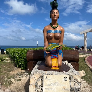 May include: A colorful statue of a woman kneeling and holding a fish. The statue is made of painted concrete and is located outdoors. The statue is wearing a blue and yellow skirt and a green headdress. The statue is in front of a sign that reads "Isla Mujeres".