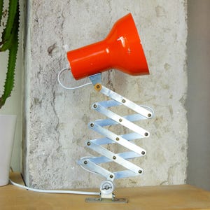Könnte beinhalten: Eine vintage Schreibtischlampe aus orangefarbenem Metall mit einem Teleskoparm. Die Lampe wird mit einer Klemme an einer Oberfläche befestigt.