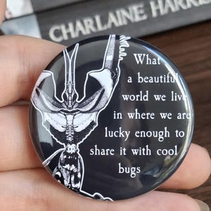 Può includere: Un bottone nero con un'illustrazione di insetto bianco e il testo "What a beautiful world we live in where we are lucky enough to share it with cool bugs."