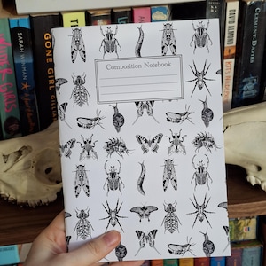 May include: A composition notebook with a white cover featuring a repeating pattern of black insect illustrations. The notebook has the words "Composition Notebook" printed on the front. The notebook is held in front of a bookshelf filled with books.
