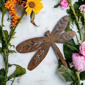 Rustic rusty metal dragonfly garden decoration with cut out v-leaf shapes in the four insect wings. Sitting on a white background with small yellow and pink flowers on greenery running up both sides of the image. Display example for yard art.