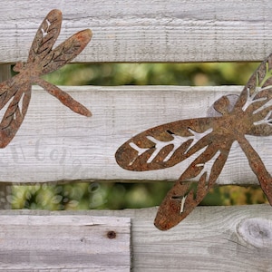 Rustic rusty metal One large and one medium dragonfly garden decorations with cut out v-leaf shapes in the four insect wings. Sitting on a weathered wooden fence with slats peeping through to greenery. The dragonflies are displayed as if flying.