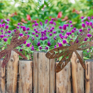 Rustic rusty metal One large and one medium dragonfly garden decorations with cut out v-leaf shapes in the four insect wings. Sitting on top of a wooden garden boarder with a bed of small purple flowers behind.